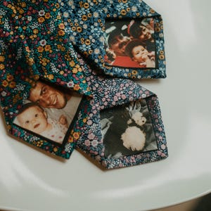 May include: Three floral patterned neckties with family photos displayed on the back. The ties have a dark blue background with small yellow, pink, and white flowers. The photos show smiling people and a baby.