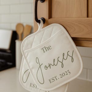 May include: Two off-white quilted pot holders with the text "The Joneses EST. 2025" printed in gray. The pot holders are hanging on a black cabinet handle in a kitchen setting.