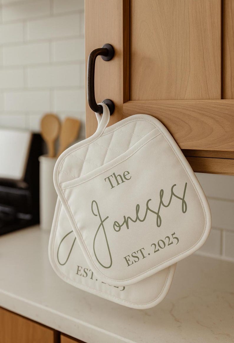 May include: Two off-white quilted pot holders with the text "The Joneses EST. 2025" printed in gray. The pot holders are hanging on a black cabinet handle in a kitchen setting.