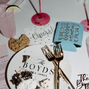 May include: A wedding table setting with a plate bearing the words "The Boyds" and remnants of cake. Gold forks, a cookie, and a blue can cooler with "Bow Down Boys" are visible. Pink glassware and a cocktail are also present.