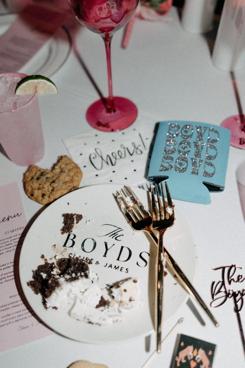 May include: A wedding table setting with a plate bearing the words "The Boyds" and remnants of cake. Gold forks, a cookie, and a blue can cooler with "Bow Down Boys" are visible. Pink glassware and a cocktail are also present.
