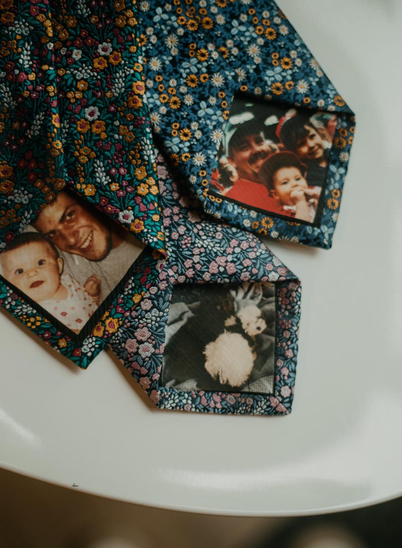 May include: Three floral patterned neckties with family photos printed on the inside. The ties have a navy blue background with a pattern of small flowers in shades of yellow, pink, and white. The photos show a baby, a family, and a dog.