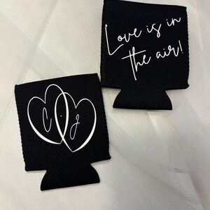 May include: Two black can coolers with white text. One cooler features intertwined hearts with the letters "C.J." inside. The other cooler says "Love is in the air!" in cursive. The coolers are on a white, textured surface.