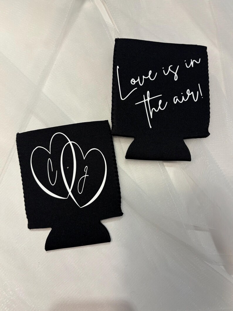 May include: Two black can coolers with white text. One cooler features intertwined hearts with the letters "C.J." inside. The other cooler says "Love is in the air!" in cursive. The coolers are on a white, textured surface.