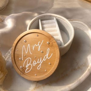 May include: A round jewellery box with a light wood lid featuring the white script text Mrs. Boyd and small white pearl accents. The base is white with a soft interior lining, including a ring roll section for organising small accessories.