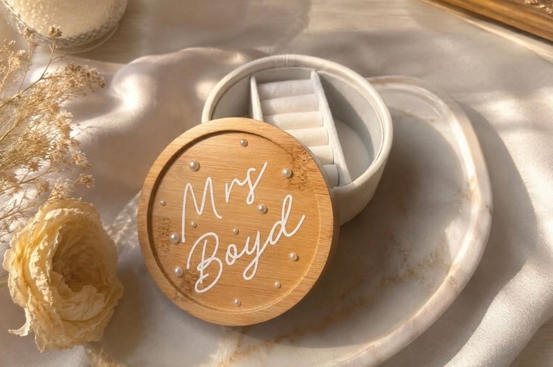 May include: A round jewellery box with a light wood lid featuring the white script text Mrs. Boyd and small white pearl accents. The base is white with a soft interior lining, including a ring roll section for organising small accessories.