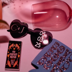May include: A selection of wedding accessories, featuring heart-shaped sunglasses with initials and a date, a matchbox reading "The Perfect Match," and a blue drinks cooler with glitter text. A pink wine glass and cocktails are also visible.