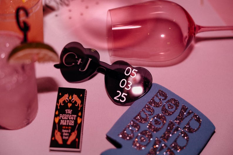 May include: A selection of wedding accessories, featuring heart-shaped sunglasses with initials and a date, a matchbox reading "The Perfect Match," and a blue drinks cooler with glitter text. A pink wine glass and cocktails are also visible.