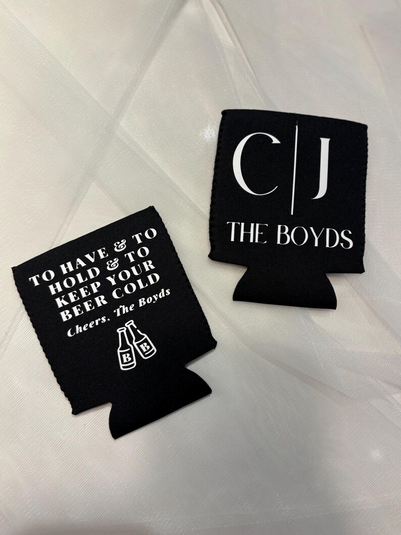 May include: Two black beer can coolers with white text. One cooler reads "TO HAVE & TO HOLD & TO KEEP YOUR BEER COLD Cheers. The Boyds" with beer bottle graphics. The other cooler reads "CJ THE BOYDS".