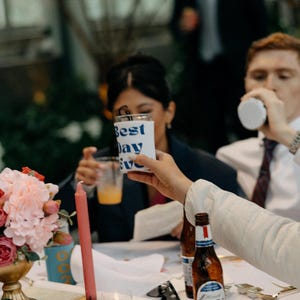 May include: A celebratory scene with a floral arrangement, candles, and drinks. A hand holds a white cup with "Best Day Ever" in blue text. Bottles of beer are on the table, and people are in the background.