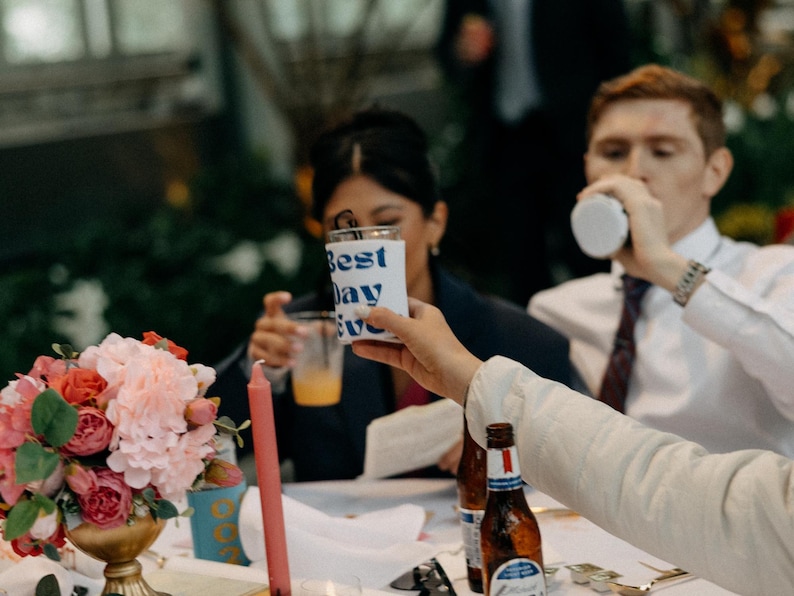 May include: A celebratory scene with a floral arrangement, candles, and drinks. A hand holds a white cup with "Best Day Ever" in blue text. Bottles of beer are on the table, and people are in the background.