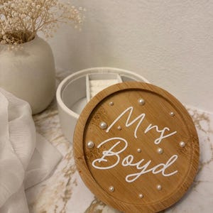May include: A round wooden jewellery box lid featuring the white script text Mrs. Boyd, accented with small white pearls embedded in the wood grain. The lid is positioned in front of a white circular jewellery container with internal dividers.