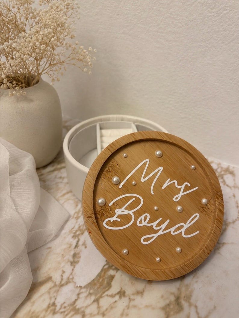May include: A round wooden jewellery box lid featuring the white script text Mrs. Boyd, accented with small white pearls embedded in the wood grain. The lid is positioned in front of a white circular jewellery container with internal dividers.