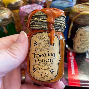 May include: A small, decorative jar labeled "Healing Potion" with a brown liquid inside. The jar has a black and gold lid with a dripping, amber-colored substance. The label includes the text "Now with +2".