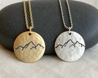 Könnte beinhalten: Zwei runde Anhänger-Halsketten, eine goldfarben und eine silberfarben, jeweils mit gehämmertem Finish und einer schwarzen Gravur eines Gebirgszuges. Der goldene Anhänger hängt an einer Gliederkette, der silberne an einer Kugelkette.