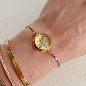 May include: A rose gold-toned bracelet featuring a small, functional circular compass charm with a gold-colored face, black cardinal direction markings, and a white needle, attached to a delicate, adjustable box chain.