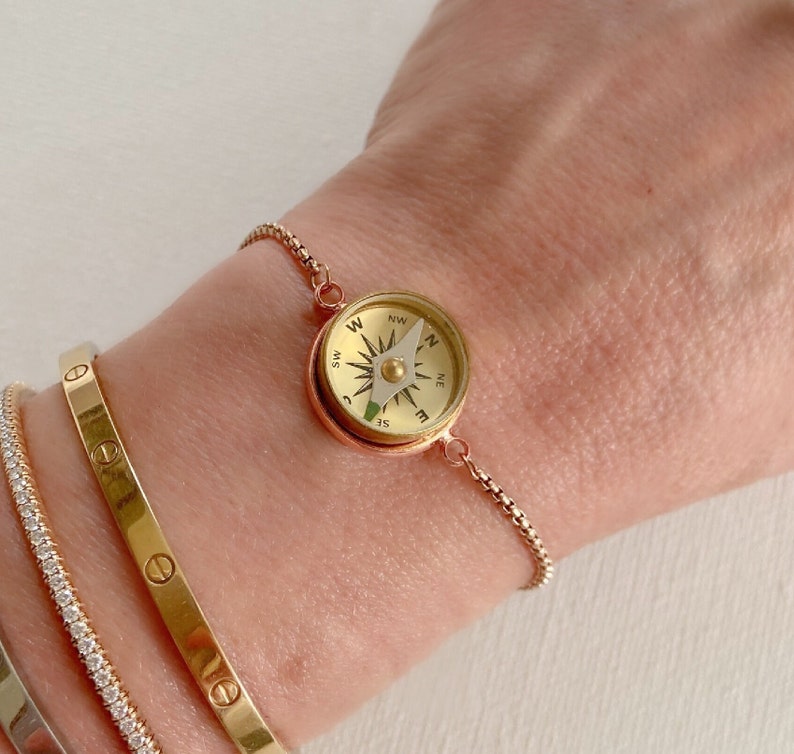 May include: A rose gold-toned bracelet featuring a small, functional circular compass charm with a gold-colored face, black cardinal direction markings, and a white needle, attached to a delicate, adjustable box chain.