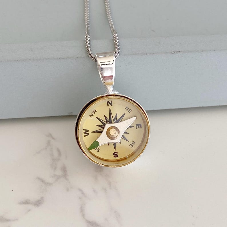 May include: Silver-toned compass pendant necklace. The compass face is cream-colored with black directional markings and a silver needle. The pendant hangs from a delicate silver chain. The compass is a classic nautical accessory.
