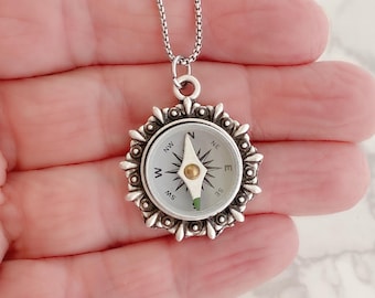 May include: A silver compass necklace with a white face and black markings. The compass has a silver bezel with a decorative design. The necklace is on a silver chain.