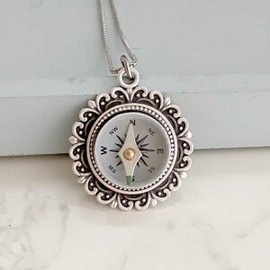 May include: Silver compass necklace with a white face and black markings. The compass features the directions north, south, east, and west. The necklace has a decorative silver frame.