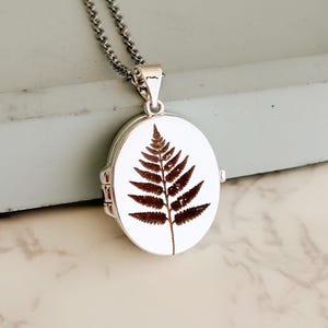 May include: Silver oval locket pendant with a detailed brown fern design on a white background. The pendant hangs from a delicate silver chain. The locket is closed and ready to wear.
