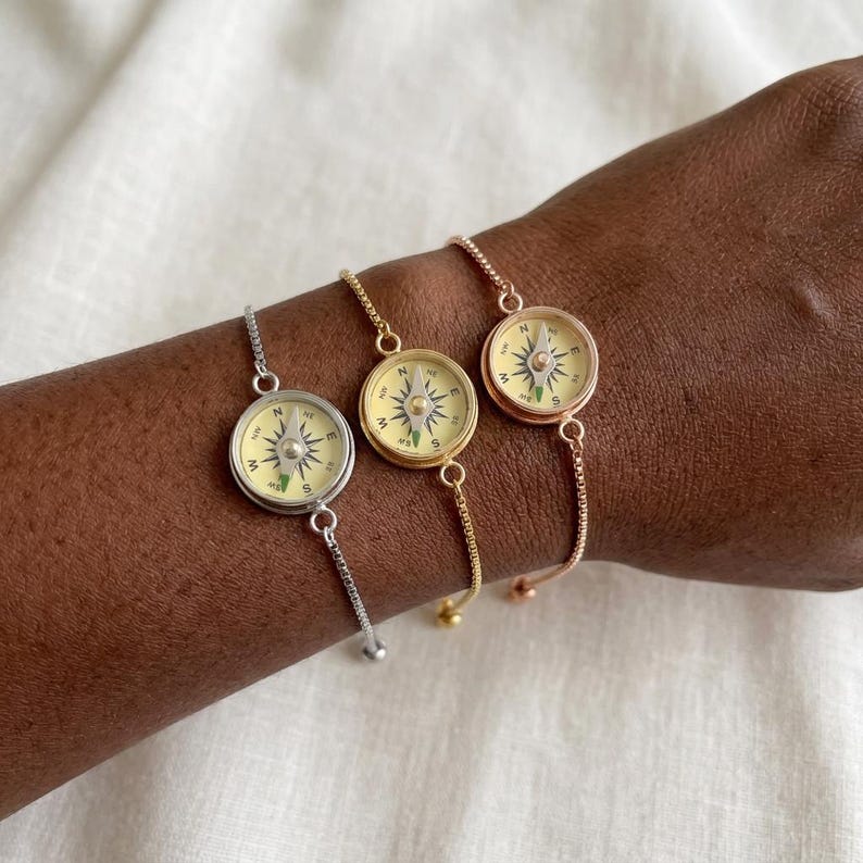 May include: Three adjustable chain bracelets featuring circular compass charms. One bracelet is silver-toned, one is gold-toned, and one is rose gold-toned. Each compass face has a cream background with black directional markings and a central needle.