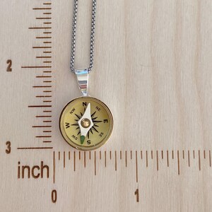 May include: A silver chain necklace with a compass pendant. The compass is gold-toned with black directional markings and a white needle. The pendant is approximately 1 inch in diameter.