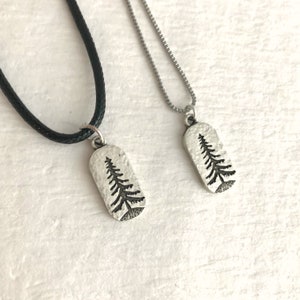 May include: Two silver-toned pendant necklaces. Each pendant is a textured, rectangular shape with a detailed black tree design. One necklace has a black cord, the other a silver chain. The pendants are displayed on a white surface.