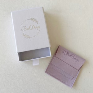 May include: An open white jewelry box with a pull-out drawer and a small, matching pouch. The box and pouch both feature the text "3 Bead Designs" in a script font. The box has a gold leaf design around the text.
