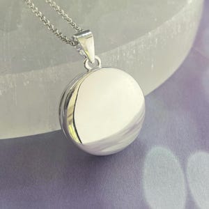 May include: A silver-colored pendant necklace featuring a round, polished charm. The charm is spherical with a smooth, reflective surface. The necklace chain is delicate and also silver. The pendant hangs from a small loop attached to the chain.