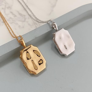 May include: Two necklaces with rectangular pendants. One is gold-toned with an abstract design, the other silver-toned with a smooth surface. Both hang from delicate chains, displayed on a light blue surface.