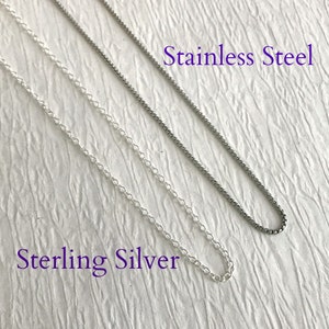 May include: Close-up of two silver-colored chain necklaces. One is a delicate, linked chain labeled "Sterling Silver." The other is a darker, textured chain labeled "Stainless Steel." Both chains are displayed against a textured, off-white background.
