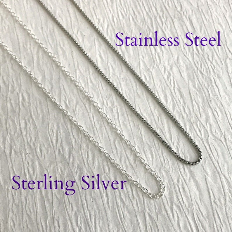 May include: Close-up of two silver-colored chain necklaces. One is a delicate, linked chain labeled "Sterling Silver." The other is a darker, textured chain labeled "Stainless Steel." Both chains are displayed against a textured, off-white background.