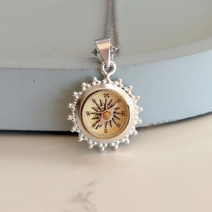 May include: Silver compass pendant necklace. The pendant features a detailed compass rose design within a circular frame. The compass has directional markings and a central needle. The necklace is suspended from a delicate silver chain, creating a classic and elegant accessory.