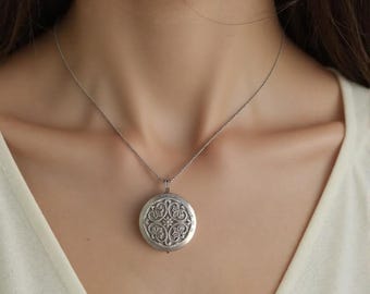 May include: A silver-toned circular locket pendant suspended on a delicate matching chain. The locket features an intricate embossed floral pattern with a central star-like detail, set against a smooth, light-coloured fabric background.