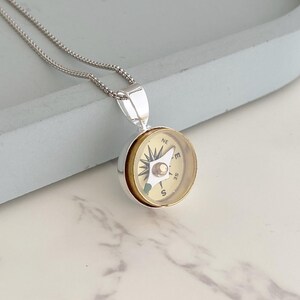 May include: Silver-toned compass pendant necklace. The compass face is gold-colored with black directional markings and a central silver-toned pointer. The necklace chain is a delicate silver bead chain.
