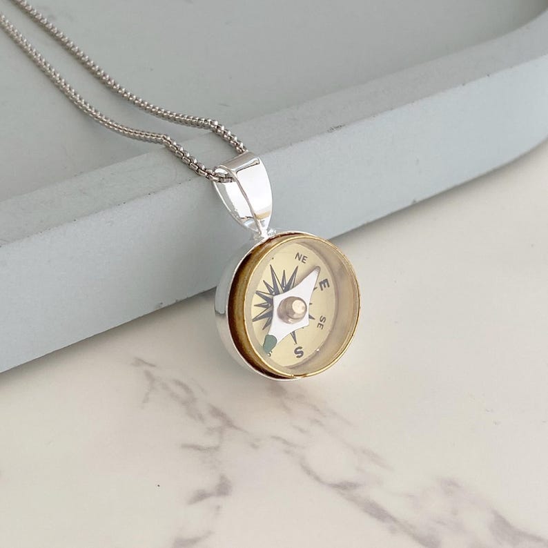 May include: Silver-toned compass pendant necklace. The compass face is gold-colored with black directional markings and a central silver-toned pointer. The necklace chain is a delicate silver bead chain.