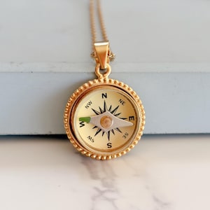 May include: Gold-colored compass pendant necklace. The compass face is cream-colored with black directional markings and a silver needle. The pendant is round with a beaded edge and hangs from a delicate gold chain.