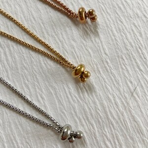 May include: Three metallic box chains in rose gold, yellow gold, and silver tones. Each chain features a sliding bead mechanism at the end, consisting of a larger rounded bead and two smaller spherical beads, allowing for adjustable length.