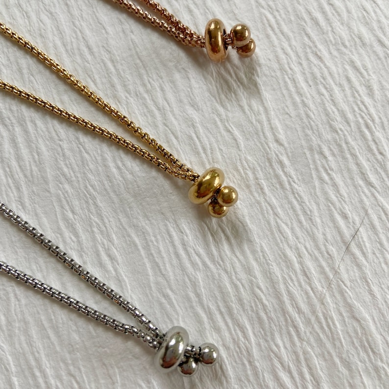 May include: Three metallic box chains in rose gold, yellow gold, and silver tones. Each chain features a sliding bead mechanism at the end, consisting of a larger rounded bead and two smaller spherical beads, allowing for adjustable length.