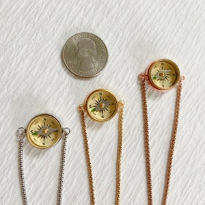May include: Three circular compass pendants in silver, gold, and rose gold tones, each attached to a matching metal chain. A United States quarter coin is included for scale, measuring approximately 0.955 inches in diameter.