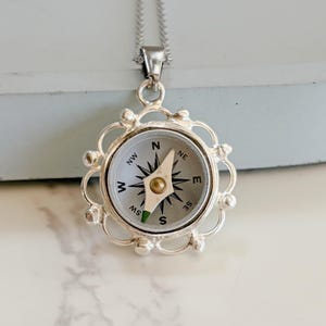 May include: Silver-toned compass pendant necklace. The compass face features directional markings and a needle. The pendant is encased in a decorative, openwork frame and hangs from a delicate chain. The compass is a classic navigational tool.