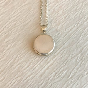 May include: A silver pendant necklace with a round, flat pendant. The pendant is attached to a delicate silver chain. The necklace is displayed on a white, textured surface. The pendant is approximately 1 inch in diameter.