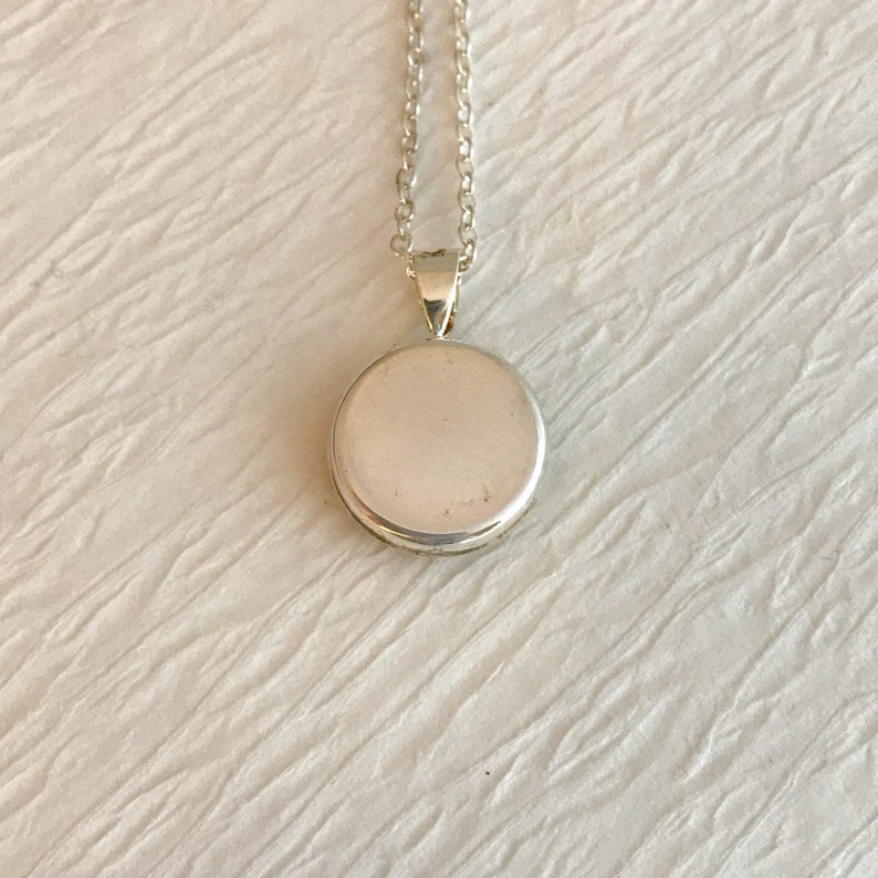 May include: A silver pendant necklace with a round, flat pendant. The pendant is attached to a delicate silver chain. The necklace is displayed on a white, textured surface. The pendant is approximately 1 inch in diameter.