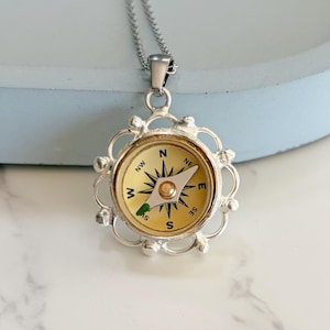 May include: Silver-toned compass pendant necklace. The compass face is cream-colored with black directional markings and a gold-colored needle. The pendant is surrounded by a decorative silver frame and hangs from a silver chain.