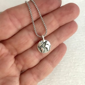 May include: A silver-colored pendant necklace featuring a round charm with a detailed world map design. The pendant hangs from a delicate chain. The necklace is shown against a light background.