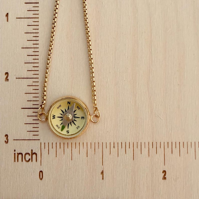 May include: A gold-toned necklace featuring a round compass pendant with a cream-colored face and black directional markings. The pendant is attached to a delicate box chain and is positioned next to a ruler showing a length of 2 inches.