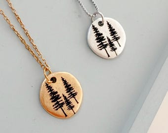 May include: Two necklaces with round metal pendants hanging from delicate chains. One pendant is gold-toned and the other is silver-toned. Each circular charm features a black engraved graphic of three pine trees of varying heights.