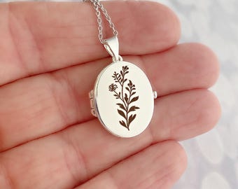 May include: An oval silver locket pendant suspended from a delicate silver chain. The front surface of the locket features a dark, etched botanical illustration of a wildflower stem with leaves and a small flower head centered on the metal.