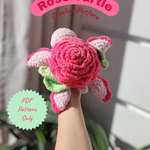 Könnte beinhalten: Eine gehäkelte Schildkröte in Form einer Rose. Die Schildkröte hat eine leuchtend rosa Rosenmitte, umgeben von rosa, grünen und weißen Blütenblättern. Das Bild enthält den Text "Rose Turtle Crochet Pattern" und "PDF Pattern Only."